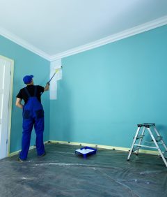 Man in a working overall is painting the wall in white color